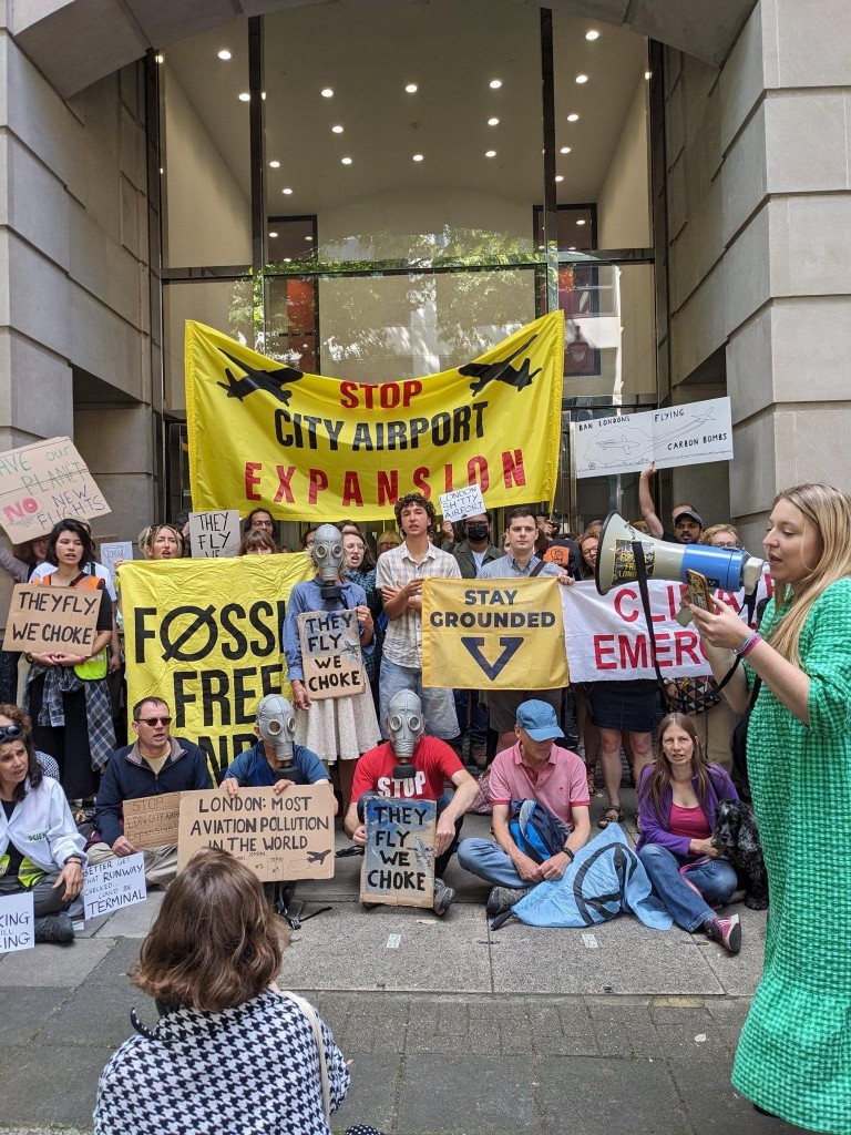 Viele Menschen, manche mit Gasmasken, mit vielen Plakaten vor einem Flughafen. Auf den Plakaten steht "Stop City Airport Expansion", "Stay Grounded", "They fly we choke"