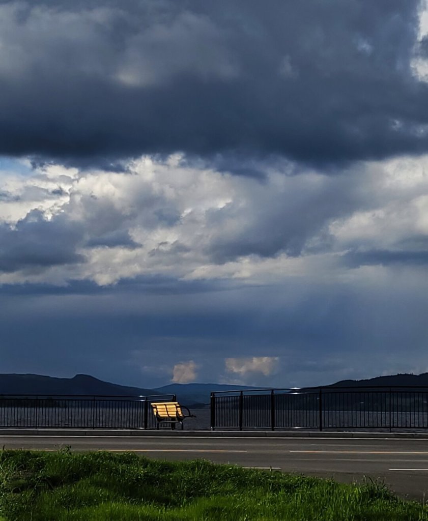 A  wooden bench on a promenade along a lake. Heavy, dark clouds are rolling in over the lake and the hills towards the bench. But they haven't reached the sun behind the bench jet and so the back of the bench is being illuminated, creating quite a contrast to the clouds