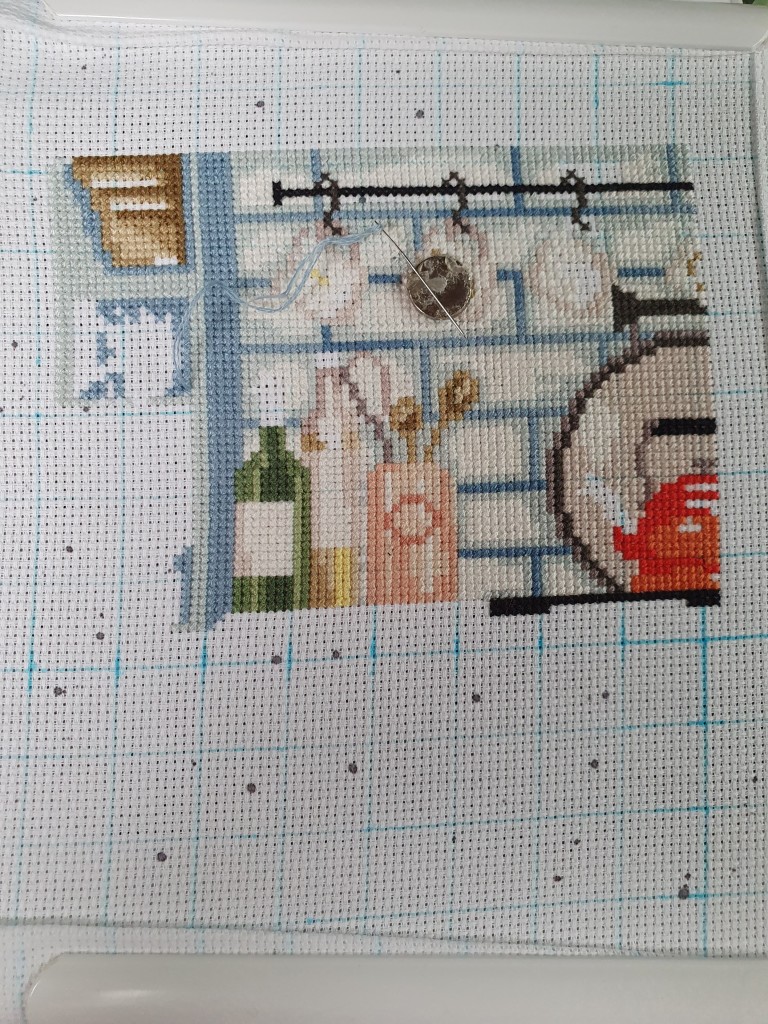 White aida with brown dots on it, with a blue hand-drawn grid on it. Cross stitches in blues, browns, greens, whites, oranges have been filled in to give a picture of tiled walks, plant, wine, oil, kettle, utensil holder & some mugs hanging. A magnet is in the centre of the fabric with a needle lying on it, which has blue thread threaded through the eye.