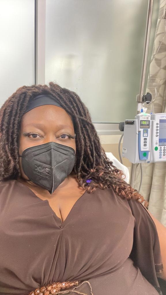 Masked selfie of me, a dark skinned Black woman  with dredlocks wearing a black face mask and a brown dress, sitting in an oncology ward with an infusion machine in the background.