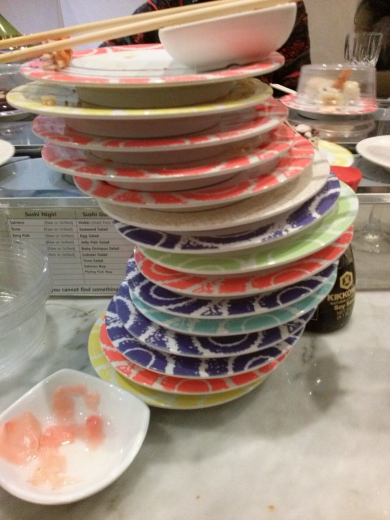 A stack of 17 sushi plates, yellow orange, blue, blue aqua blue, orange, green blue. No idea orange, orange, yellow, orange they're stacked towards the right and then they come back towards the left. On top of the stack is little ginger bowl and a pair of chopsticks