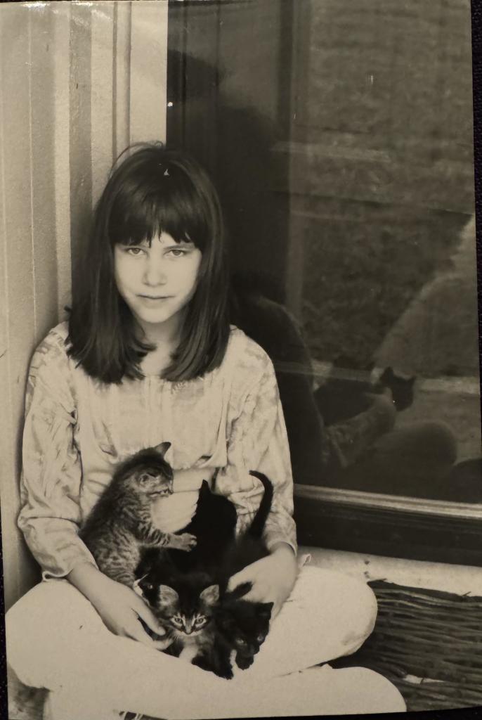 A girl sitting against a wall holding several kittens in her lap, all in black and white.