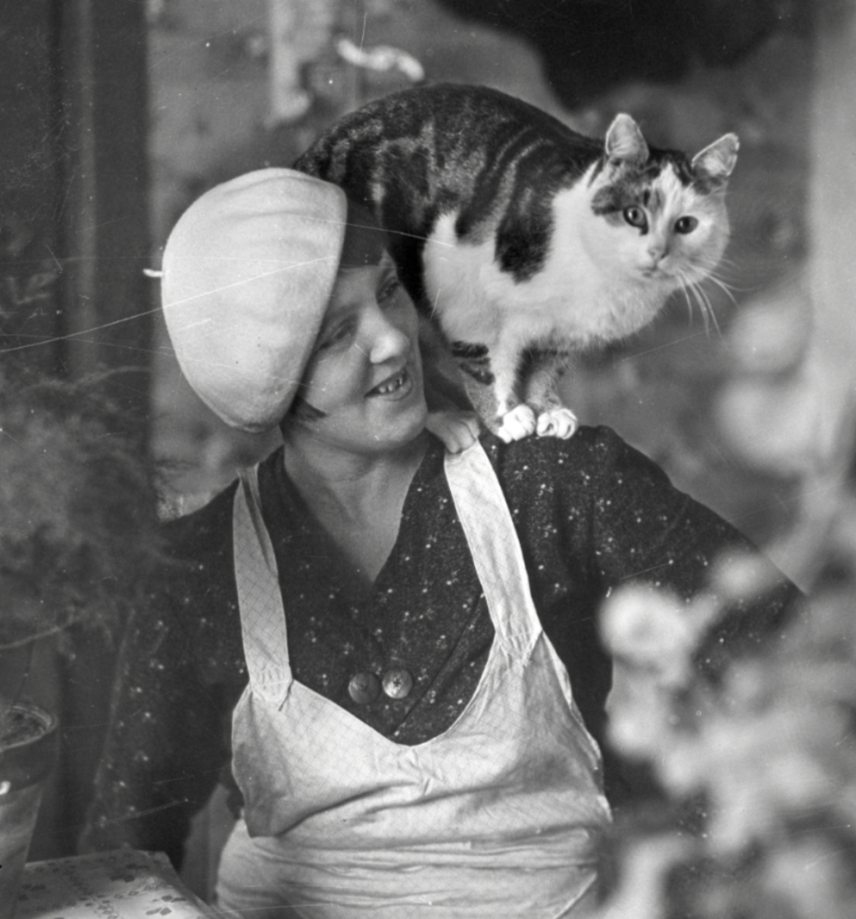 Black and white photo of a white woman in a light beret, dark dress with tiny floral print, and a a light colored apron, sitting at a kitchen table. She's smiling and looking to the left, while on her shoulder is perched a chubby shorthaired white and tabby cat that gazes confidently directly at the camera.