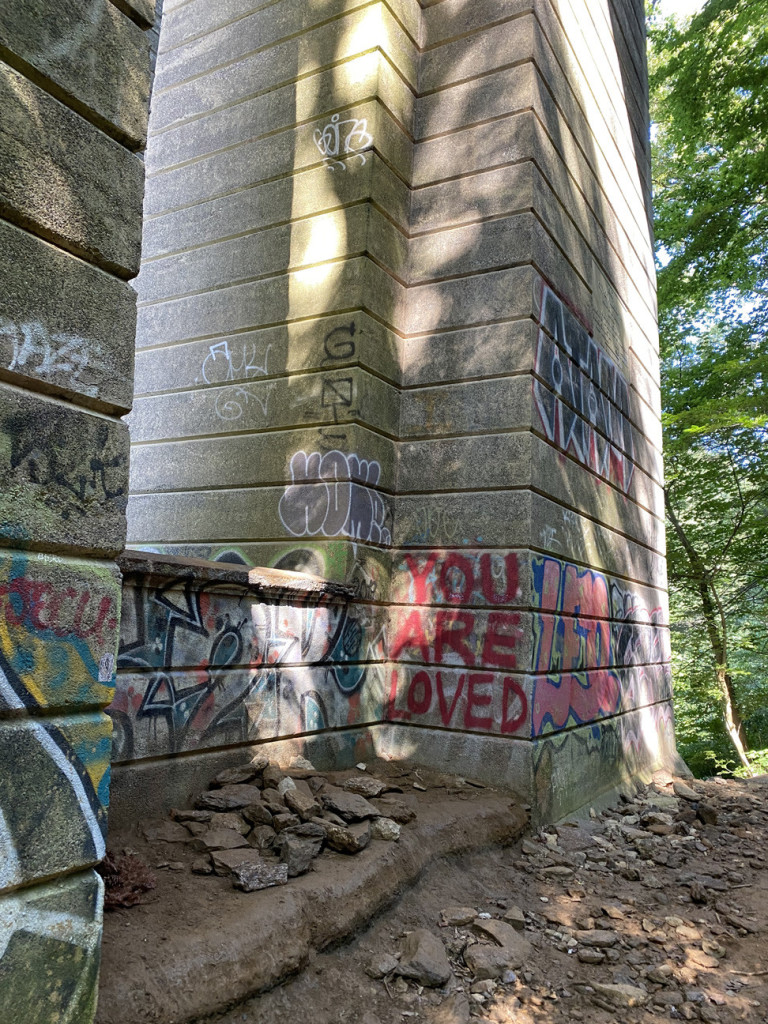 Supports for a bridge, partially lit through the trees, covered with various painted graffitti.  Most legible in the lower center is red lettering that reads You Are Loved.