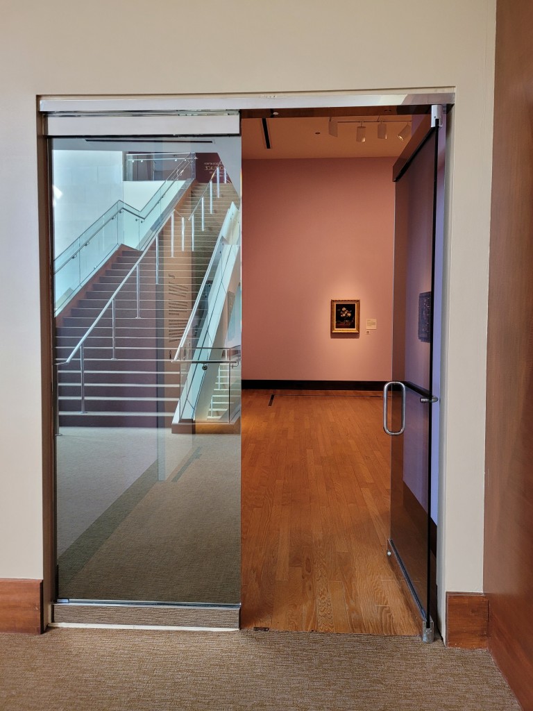 Two glass doors into a museum gallery. One is closed and shows thee reflection of the atrium stairway. The other is open and a single painting is seen on the far wall.