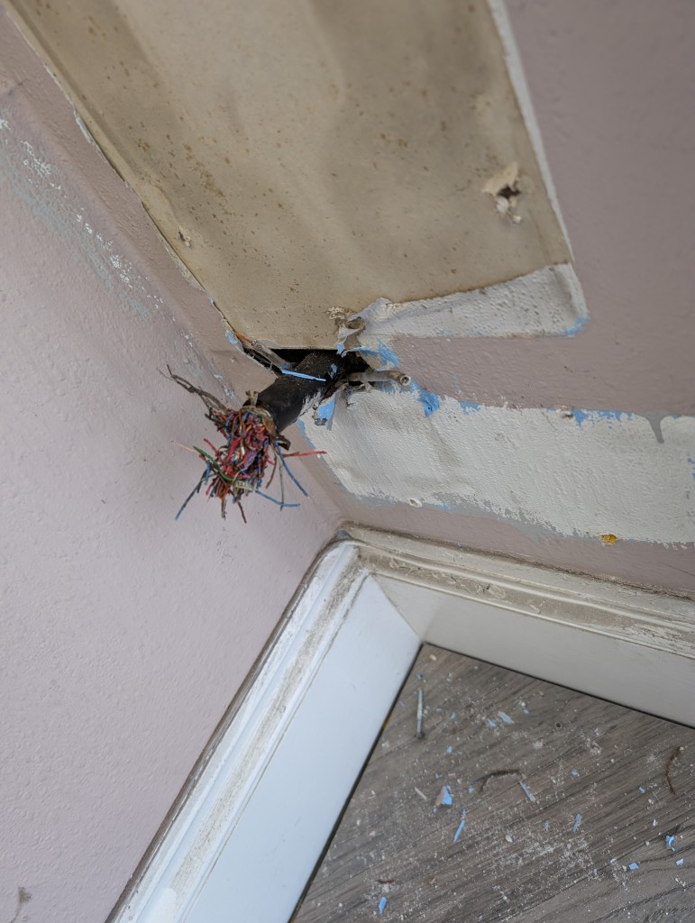 A photograph of a partly dismantled office, through the wall pokes a large black cable with a bundle of smaller wires within, and two small telephone cables.. all of which are cut.