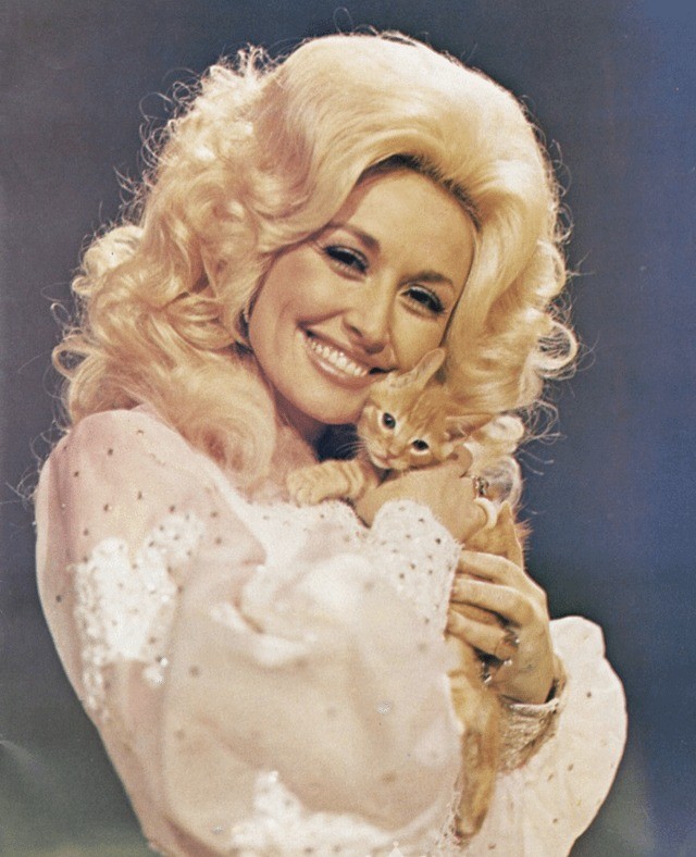 Dolly Parton, in a dress with ploofy long sleeves, holding a kitten