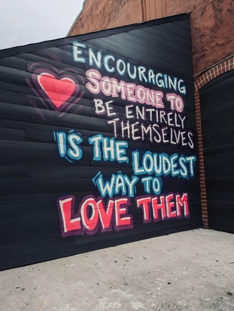 Street art mural on a dark wall featuring an uplifting message in colorful text. The message reads, 'Encouraging someone to be entirely themselves is the loudest way to love them,' with 'LOVE' highlighted in large, bold letters. A red heart is painted beside the word 'Encouraging,' adding warmth to the message. The text is done in various shades of blue, white, and pink, creating a vibrant and positive statement about acceptance and encouragement.