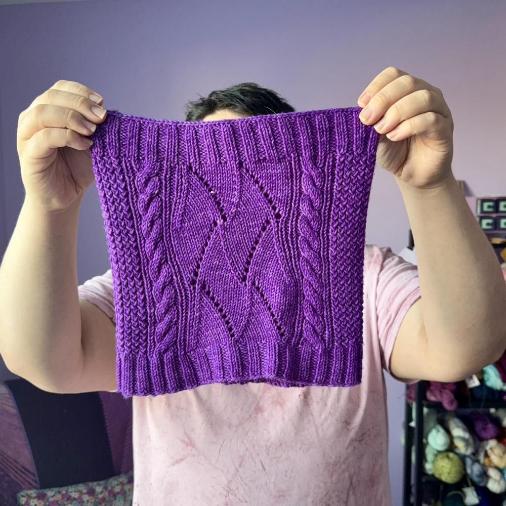 Holding the purple cowl up to the camera. In the center of the cowl is a wavy lace pattern bordered on each side by vertical cables. In either side of the cables is a textured slip stitched design