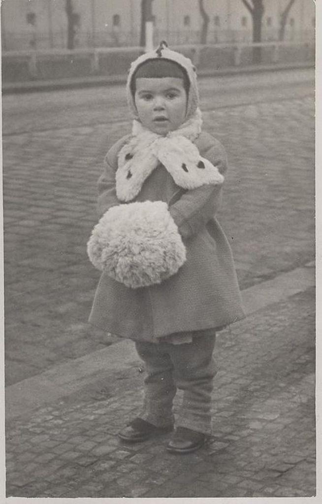 A vintage black and white photo of a young child standing on a street, wearing a coat with a large fluffy collar and a matching hat, holding a large pom-pom.