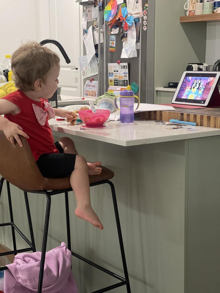 Babby, sitting on a high chair, eating Weet-Bix and watching Bluey. 