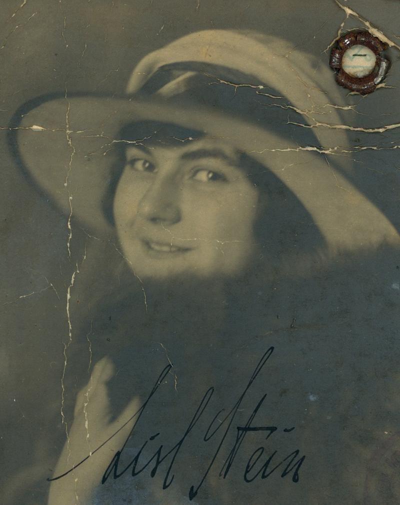 Vintage scratched photograph of a woman in a wide-brimmed hat, featuring a gentle smile. The image includes an autograph and a small, damaged area that obscures part of the face.