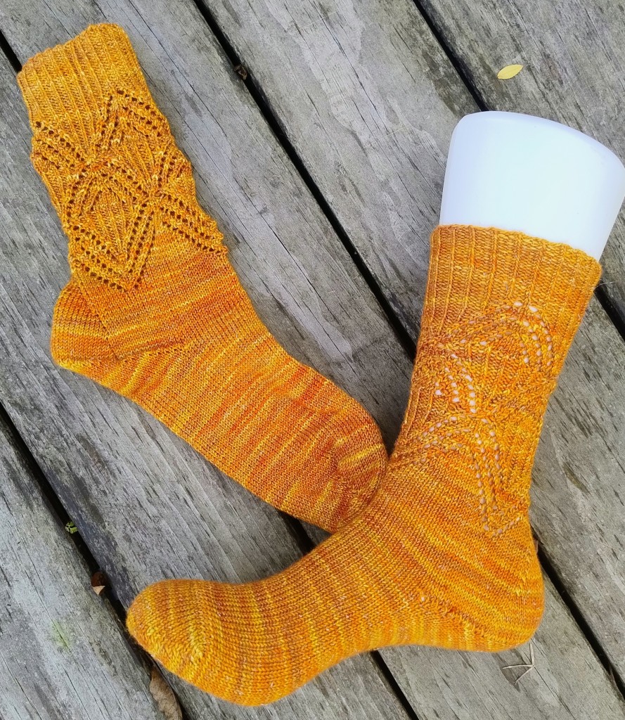 
A pair of hand-knit socks in a vibrant orange-golden hue, showcasing a delicate lace pattern and shimmering with subtle glitter, rest on a weathered wooden deck. One sock is modeled on a mannequin foot, highlighting the intricate design.
