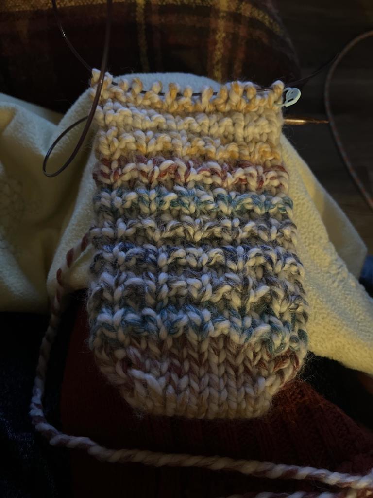 A thick looking bottom portion of a sock with no heel or ankle yet. It is made with thick yarn that changes between several retro colors including yellow, orange, blue, gray, and cream. It has a bumpy pattern