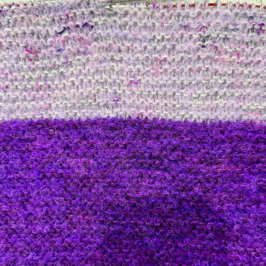 Close up of Knitted garter stitch fabric from a shawl in progress. There are two sections of colors. The bottom half is a purple yarn with fuzzy neon purple yarn to highlight, the top half is a gray  yarn with purple and pink variegations accented by fuzzy pink yarn