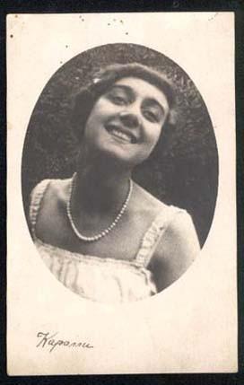 Black and white photo, 1910s. She has short, dark, straight hair. She is wearing a sleeveless white dress and a pearl necklace