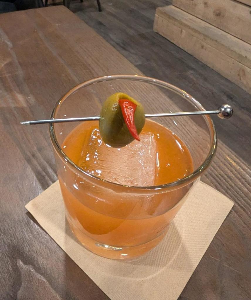 Picture of a drink on a table with a silver metal toothpick over the top that has an olive and pepper on it