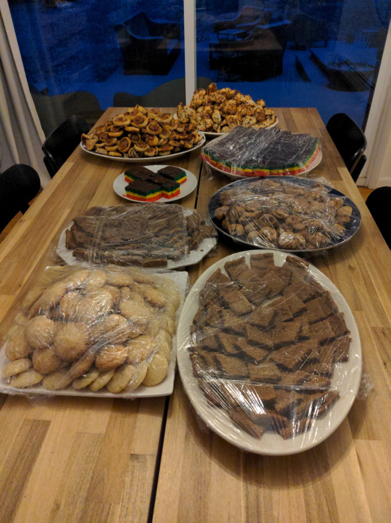 A table with multiple trays heaped with freshly baked holiday cookies: coconut macaroons, Venetian tri-color layer,  cherries jubilee, ginger hermit bars, almond, and rugelach pinwheels. In the background you can see through sliding glass doors snow settling on the deck.