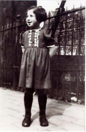 A vintage black-and-white photograph of a young girl in a patterned dress, smiling and standing in front of a metal fence.