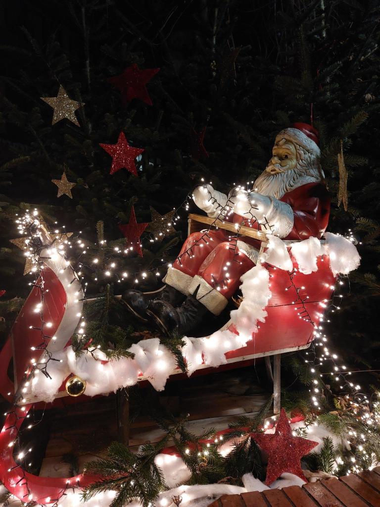 Ein roter Schlitten mit Lichterkette. Auf dem Sitz eine große Weihnachtsmannfigur. Vor dem Schlitten liegen Geschenke.