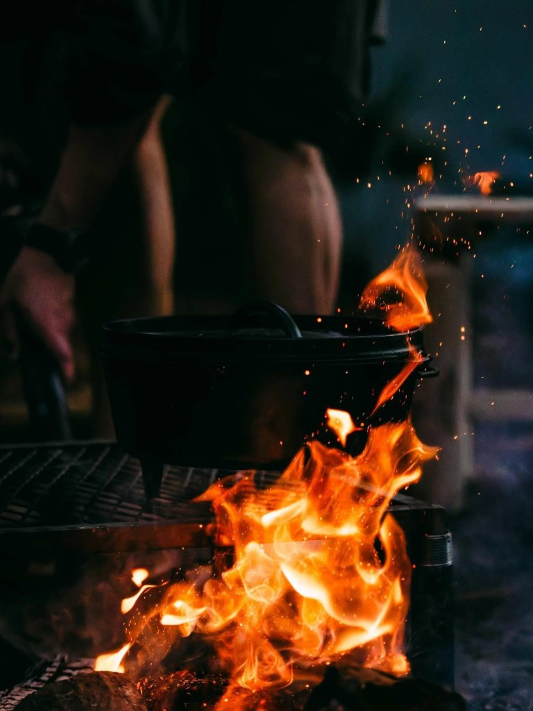 Das Bild zeigt einen gusseisernen Topf, der über einem offenen Feuer steht. Die Flammen schlagen hoch und umspielen den Topf, während im Hintergrund eine Person unscharf zu erkennen ist, die vermutlich das Feuer schürt oder den Topf bedient.Es könnte sich um eine Szene beim Camping oder einer Outdoor-Küche handeln, wo traditionell über offenem Feuer gekocht wird.