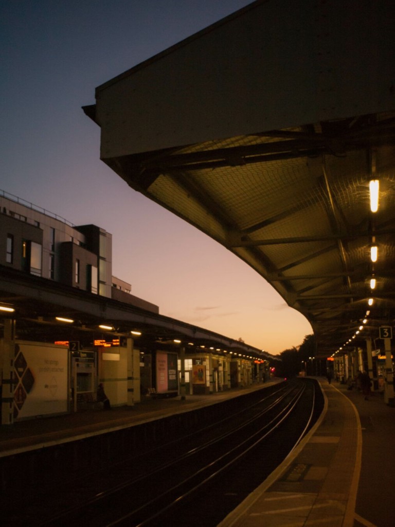 Evening photo in which two railway tracks run between canopied platforms, the curve that they all follow is accentuated by the wide angle of the lens. The picture is taken from the platform on the right, where the canopy swings from the top left down to the bottom middle, under which runs a line of long lights. This platform is mirrored on the on the other side of the tracks, which gleam in the light from the violet-to-orange-pink sky above. Behind the far platform rises a blicky building, also touched by the sunset light.