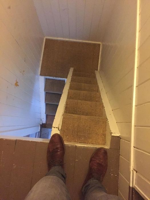 Photo looking down at the some rather narrow stairs going at the end of a rather narrow hallway. It almost look more like stairs that were made for cats in the way it's been divided and turns back on itself without guard rails. Make sure to watch your step!