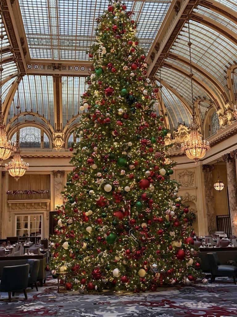 A large Christmas tree adorned with red, green, and gold ornaments stands beneath a grand, arched glass ceiling. 