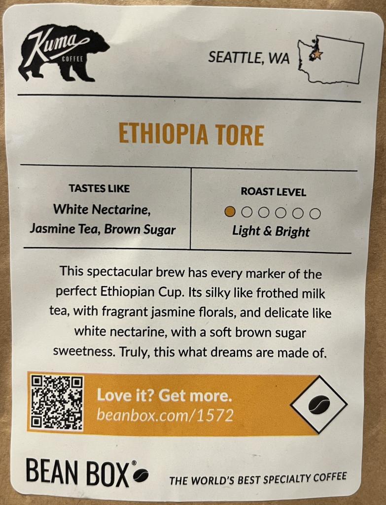 The label from a packet of coffee beans. At the top left  is a black logo featuring a bear with “Kumas COFFEE” written in white inside the bear. 

Top right is a small outline of Washington with “SEATTLE, WA” beside it. 

Below is the coffee information: 

ETHIOPIA TORE
TASTES LIKE
White Nectarine,
Jasmine Tea, Brown Sugar

ROAST LEVEL
Light & Bright (in a row of six dots only is colored in orange)

This spectacular brew has every marker of the perfect Ethiopian Cup. It’s silky like frothed milk tea, with fragrant jasmine florals, and delicate like white nectarine, with a soft brown sugar sweetness. Truly, this what dreams are made of.

In an orange bar, there is a QR code to the left, then “Love it? Get more. beanbox.com/1572” and a black coffee bean inside a black diamond outline at the right end of the bar.  

BEAN BOX (black coffee bean)
THE WORLD'S BEST SPECIALTY COFFEE