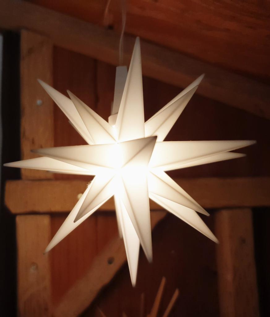 Ein Herrnhuter Weihnachtsstern leuchtet weiß vor einer Wand, die mit Holzleisten verkleidet ist.