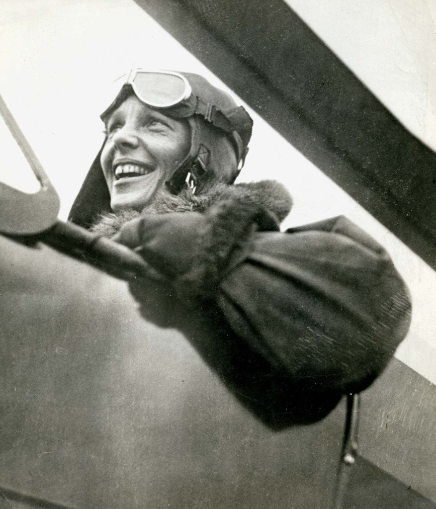 Still image. Black and white photo of aviator Amelia Earhart in an open-canopy plane, smiling broadly, wearing a winter coat, leather cap, and goggles on forehead. 