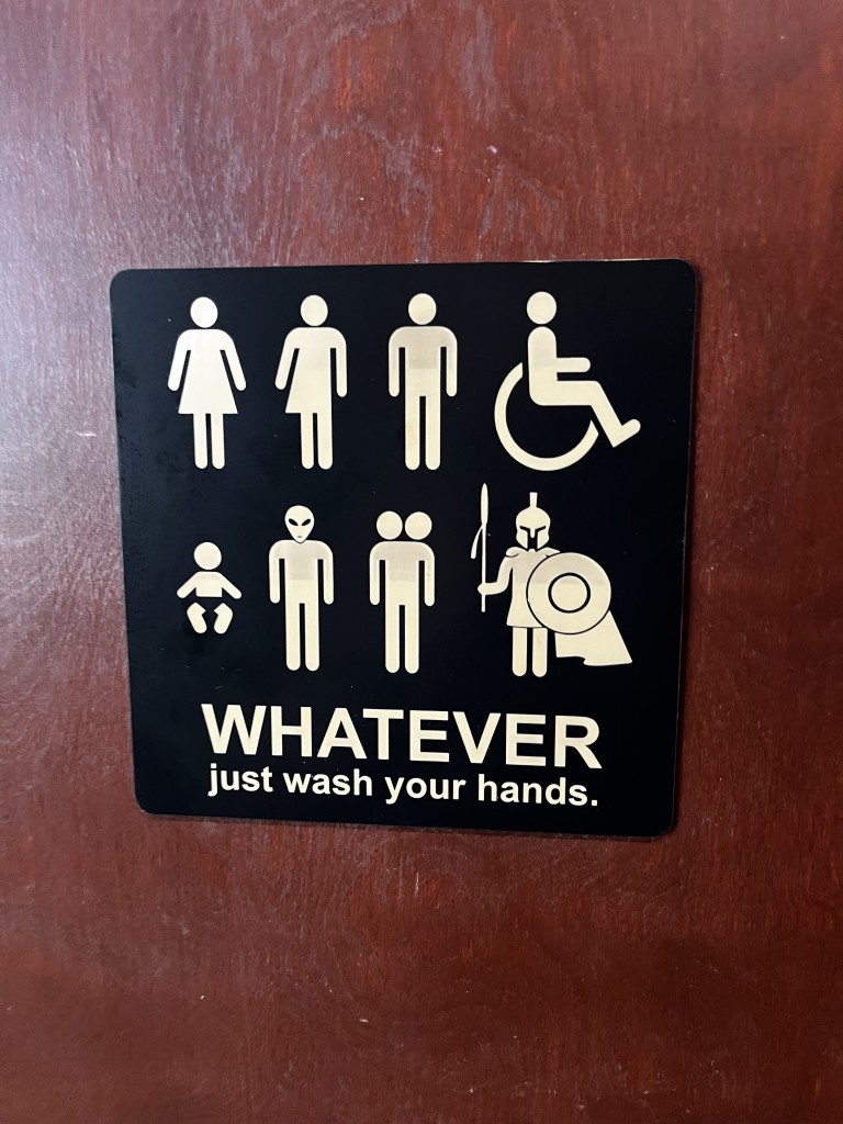 A bathroom sign, and instead of a man or woman symbol, it has a man, a woman, a person who is half of each, a person in a wheelchair, a baby, an alien, a person with two heads, and a Spartan holding a spear and shield. It says "WHATEVER. Just wash your hands."