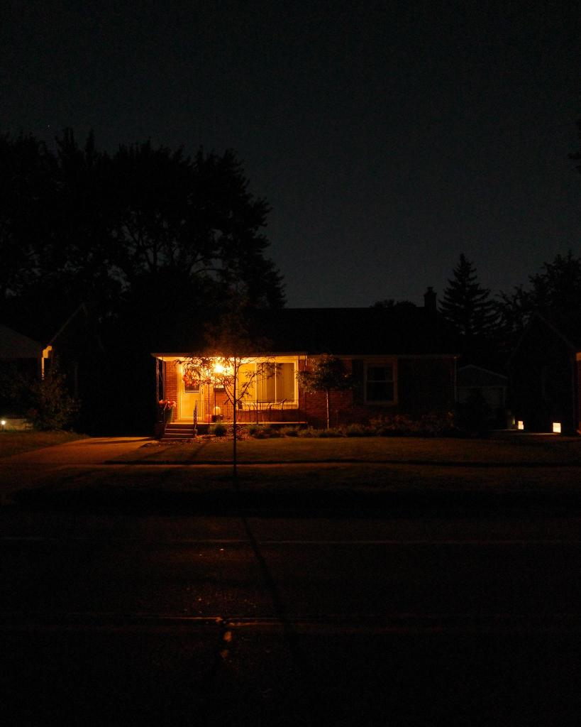 Color photograph looking across a darkened street at night at a small brick house with a porch light shining a bright golden color and some Halloween decorations on the porch, trees looming behind the house silhouetted against a clear night sky glowing silver from city lights