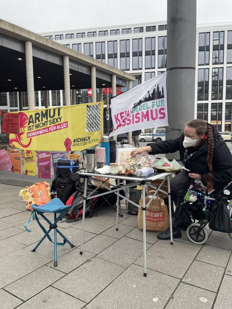 Eine Frau im Rollstuhl arrangiert Essen auf einem Tisch in einer Außeneinrichtung mit Bannern gegen Armut und Kapitalismus in der Nähe.