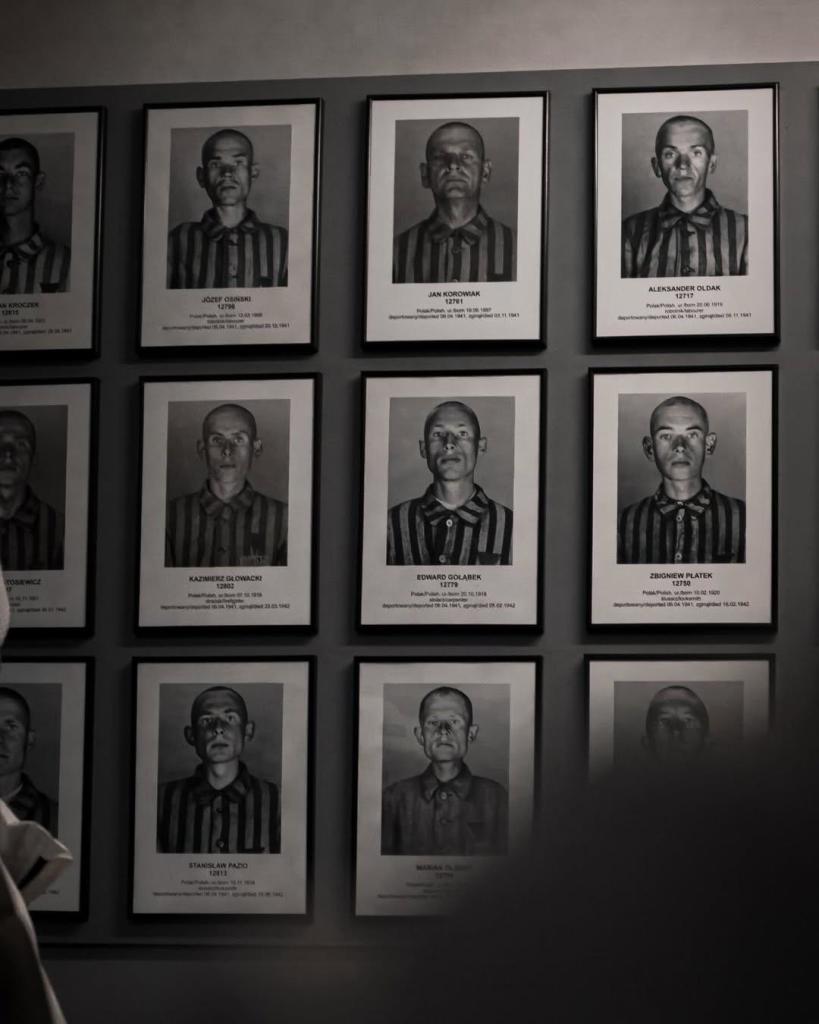 A wall displaying a grid of framed portraits of prisoners from a concentration camp. Each frame contains a black-and-white photograph of a prisoner wearing striped uniforms, along with their names, assigned numbers, dates of birth, and other identifying information. 