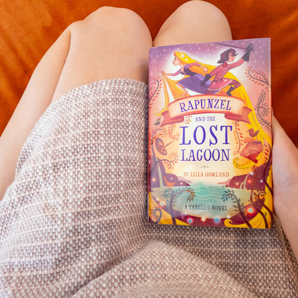 Photo of Lety sitting on an orange sofa with the copy of “Rapunzel and the Lost Lagoon” by Leila Howland she borrowed from the library on her lap. From top to bottom, she is seen wearing a beige knitted skirt. The photo is cropped in on Lety's lap on the couch, the book on her right hand, which is on her right thigh.