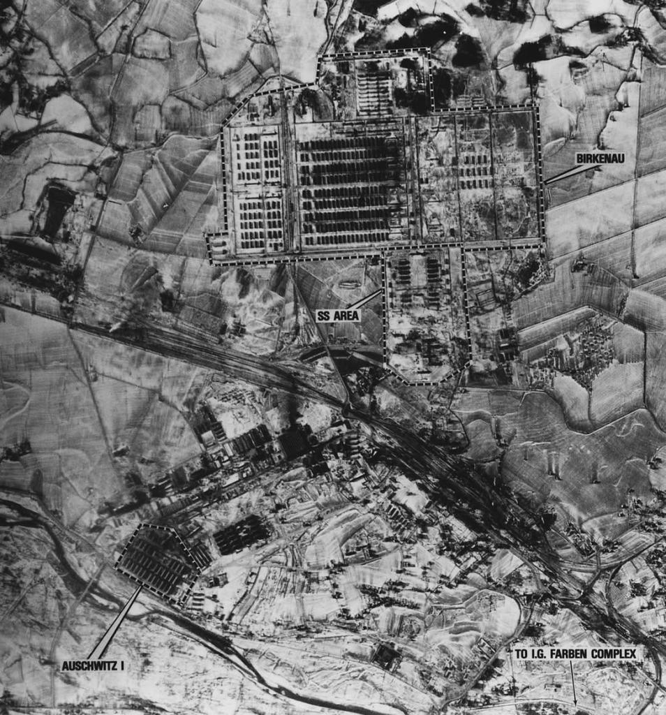 A black-and-white aerial photograph depicting Auschwitz-Birkenau concentration camp during World War II. The image reveals the vast layout of the camp, with clearly labeled sections, including "Birkenau," "SS Area," and "Auschwitz I." The arrangement of barracks, railway lines, and surrounding fields is starkly visible, showcasing the systematic and expansive structure of the site. The photograph also marks the railway connection leading to the IG Farben complex