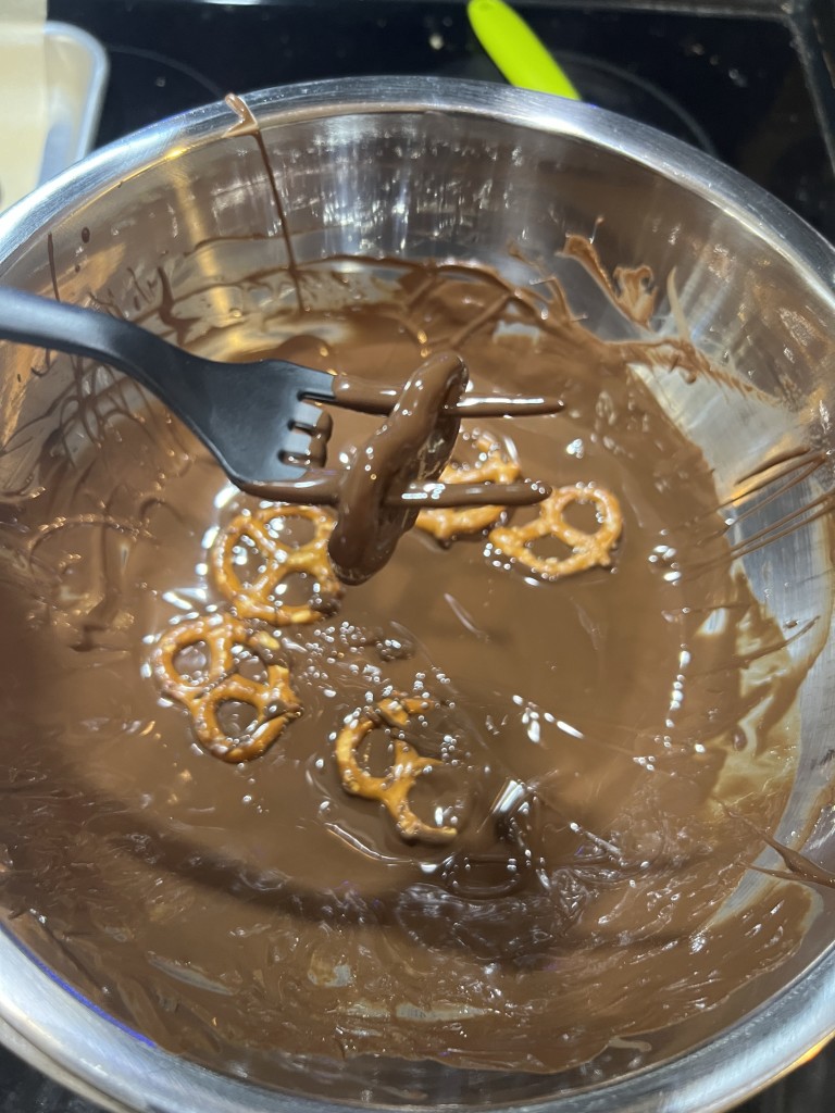 Photo of a double boiler bowl full of melted chocolate and a 2-tine fork with a chocolate covered pretzel hanging off it. Some other pretzels are floating in melted chocolate. 