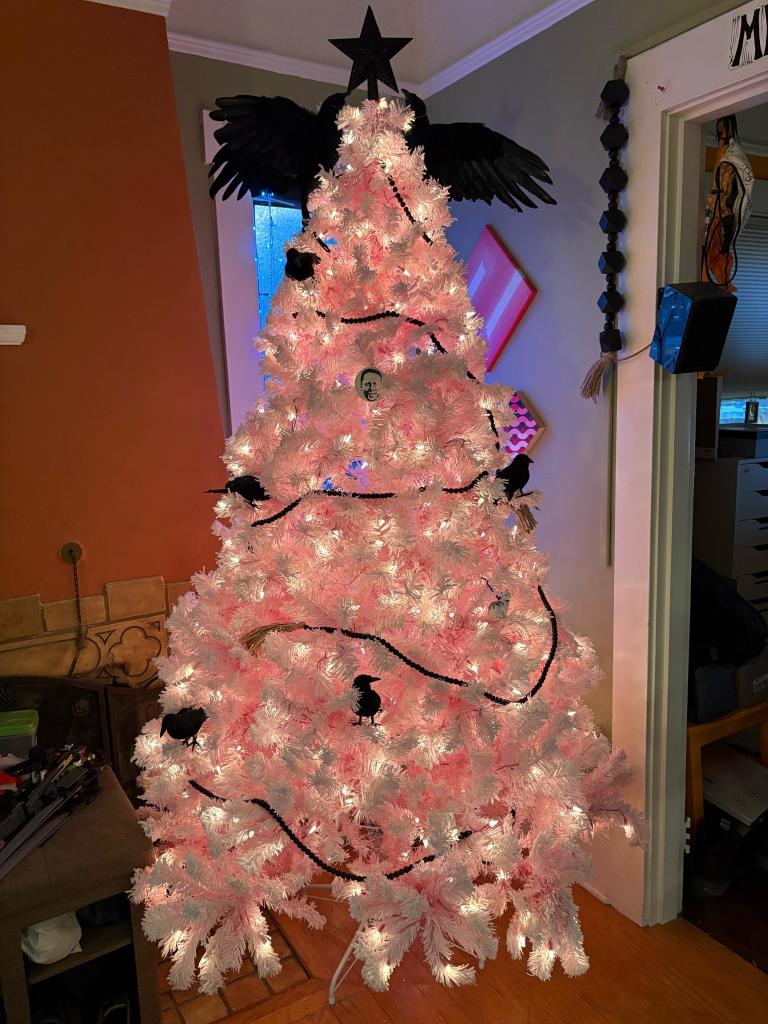 Pink artificial Christmas tree flocked with fake snow and draped with black garlands, several small corvids, and topped with a black star flanked on each side by crows with wings outstretched 