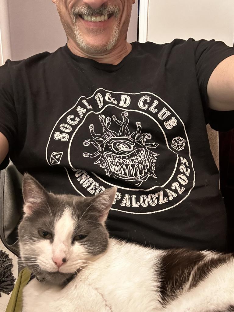 Grey and white cat in my lap chilling. I’m giggling because of her loud purr. I’m wearing a black shirt with a Beholder on it.