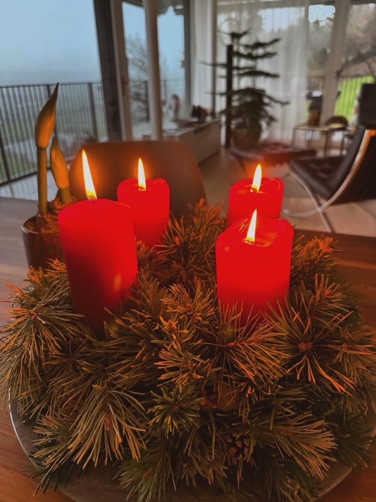 Ein dekoratives Arrangement mit vier brennenden roten Kerzen, umgeben von Tannenzweigen, die auf einem Tisch stehen. Der Hintergrund zeigt einen nebligen Blick durch ein Fenster mit sanftem Licht.