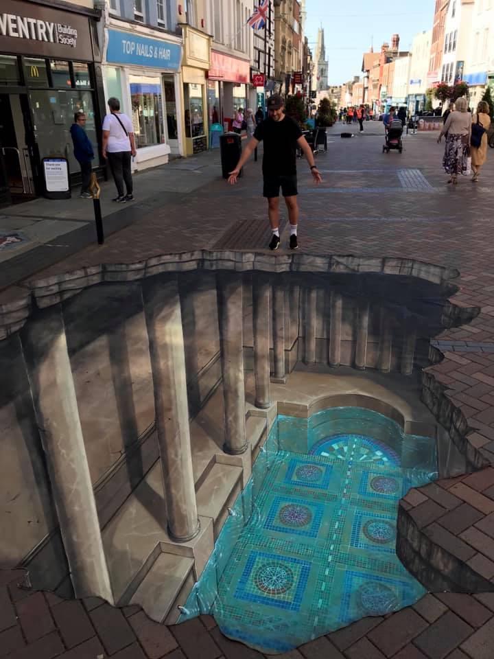 A detailed 3D street art illusion by Joe and Max showcasing a hidden Roman-style temple beneath a bustling pedestrian street. The artwork features grand stone columns and vibrant blue mosaic tiles, creating the appearance of an underground chamber. A man stands at the edge of the illusion, appearing as though he is balancing above the chasm. The realistic details blend seamlessly with the urban environment, captivating passersby.