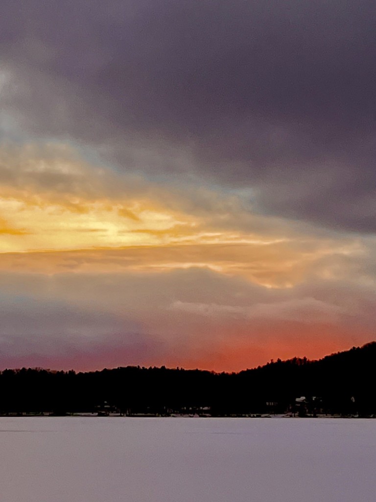 A winter sunset over a frozen lake, with dark clouds and vibrant orange and pink hues illuminating the sky above a tree-lined horizon.