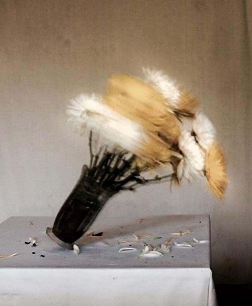 Photography. A color photo of a falling flower vase. The photo shows a dark glass vase with white and yellow flowers on a table with a white tablecloth. The vase is in motion, the image is blurred due to the rapid movement, individual petals rain down on the table while the vase tilts to the right. A new interpretation of a floral still life in a fast-paced form.