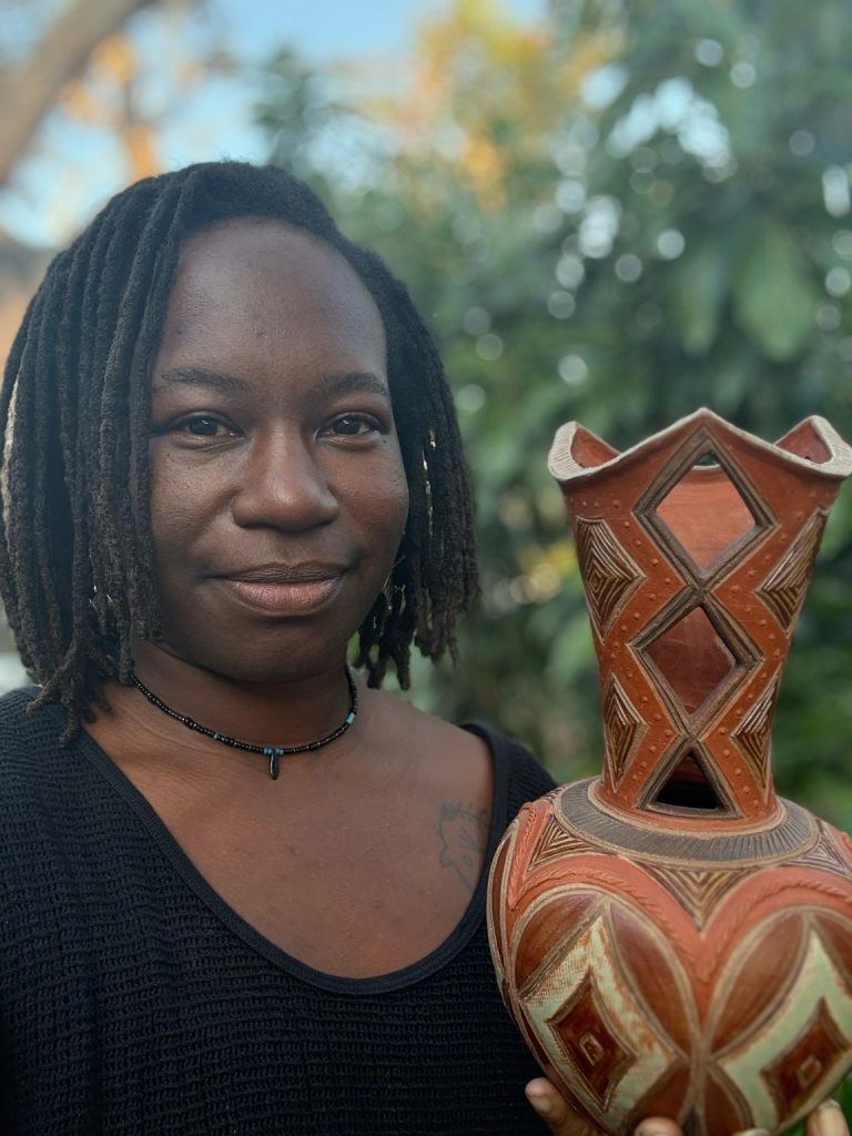 Nigerian-American studio potter Osa Atoe (a black woman with shoulder length locs)  holding an elaborate ceramic vase she made, painted in earth tones, with geometric patterns. The background is a blur of tropical foliage. 