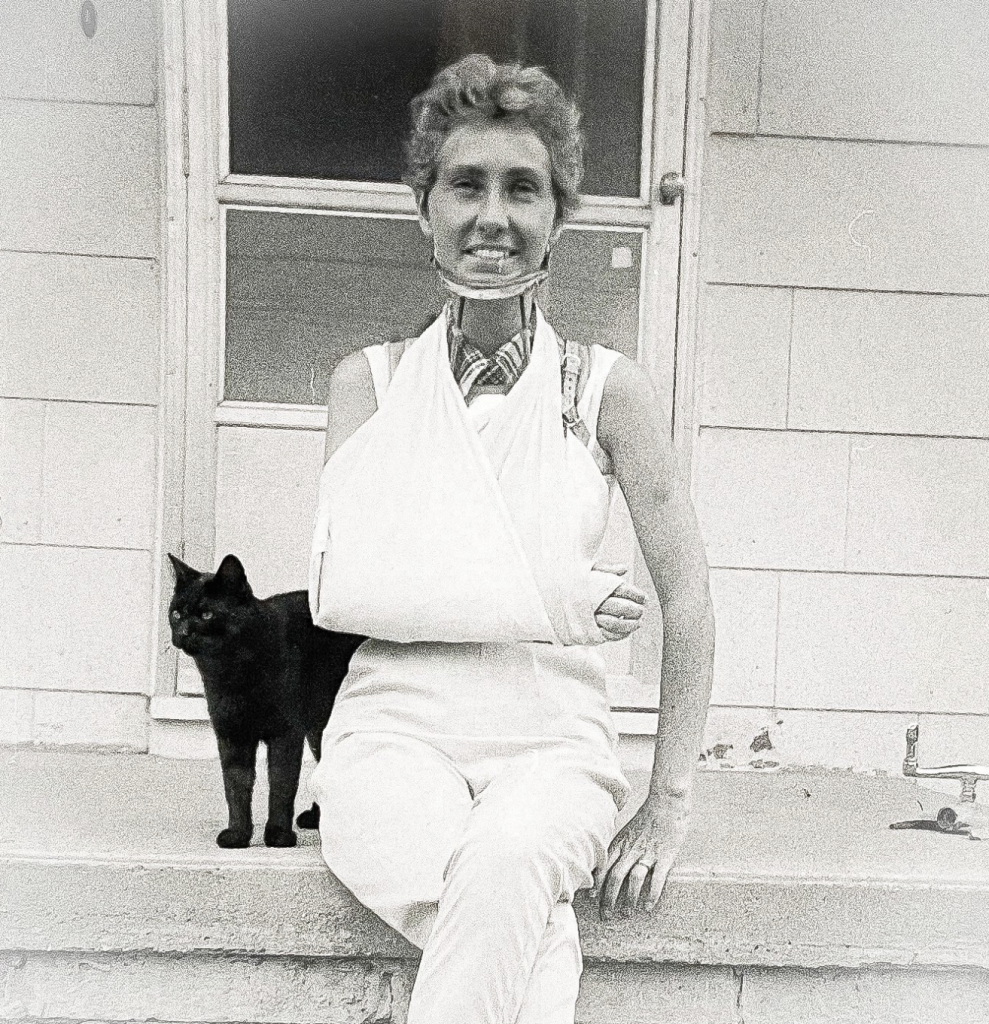 Black and white photo of a smiling white woman sitting outside on a sunny house stoop, wearing a cast and sling on her right arm and cervical collar. A happy little shorthaired black cat rubs up beside her.