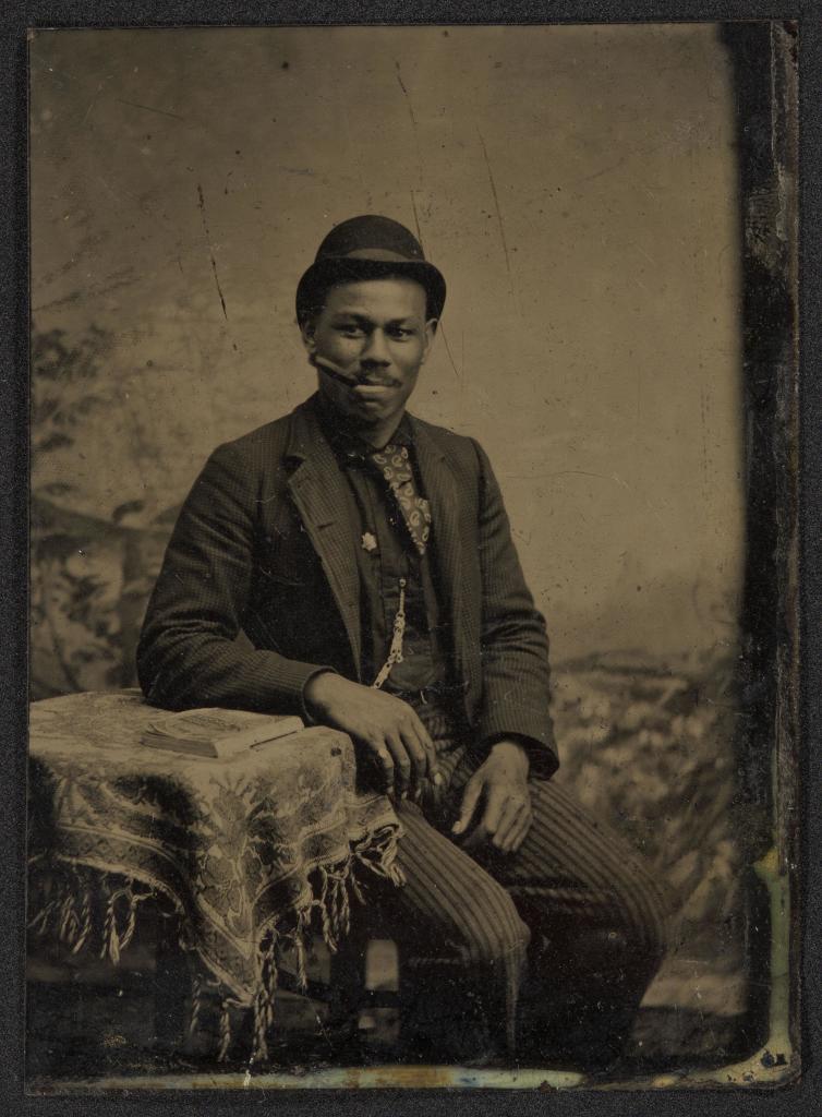 Studio portrait of man sitting with arm resting on table with book, cigar in mouth, circa 1860s-1880s 