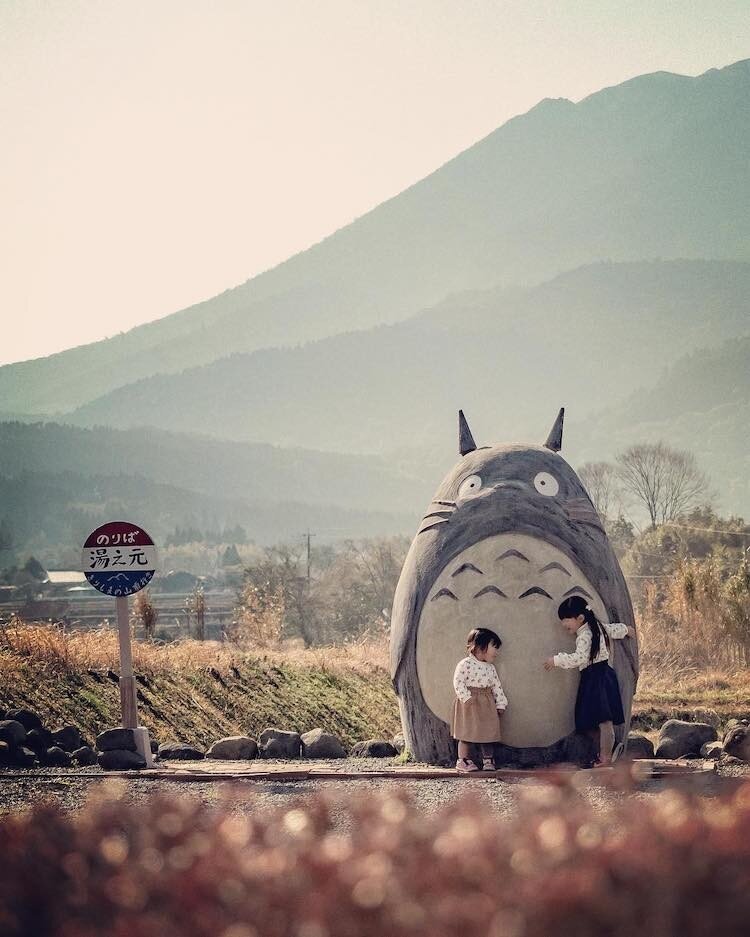 Two young children interact with a life-size Totoro sculpture from Studio Ghibli's My Neighbor Totoro in Takaharu, Japan. The Totoro stands tall with its round belly and signature features next to a red and blue bus stop sign. The backdrop features a hazy mountain and rural fields, creating a peaceful and whimsical atmosphere. The children appear to be playing and exploring near Totoro, adding a heartwarming touch to the scene.