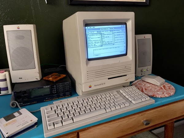 A Macintosh SE/30 sitting on a midcentury desk with the top painted a real color. The Mac is running Macstodon 0.3. Other accoutrements on the desk are a stack of floppy disks, some MiniDiscs, AppleDesign powered speakers, and a Roland SC-88Pro MIDI module.