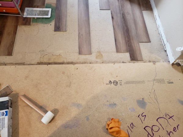 View of flooring in progress: at the very top of the photo are fingers of laminate planks of various offsets reaching toward the center

Just above center is a border between new and legacy subflooring; the new stuff is the lower half of the picture

Also shown is a rubber mallet, an orange rag, and a partial sentence scrawled in purple by an eleven year old